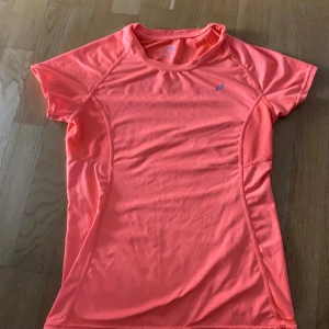 Korallrosa träningst-shirt Pro Touch - Sportig korallrosa t-shirt från Pro Touch med korta ärmar och meshpaneler på sidorna för extra ventilation. Tillverkad i lätt och snabbtorkande syntetmaterial, perfekt för träning och aktiva dagar. Snygg passform och diskret logga på bröstet.