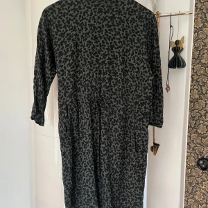 Mörkgrön blommig klänning Indiska S - Mysig klänning från Indiska i mörkgrönt med svart blommigt mönster. Klänningen har trekvartsärm och knytband i midjan för en snygg passform. Perfekt för dig som gillar en avslappnad men ändå cool stil.