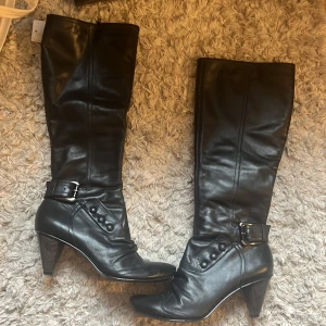 Svarta höga stövlar med klack - Snygga svarta stövlar i skinn med hög klack och spetsig tå. Dekorativa knappar och spänne vid ankeln ger en cool touch. Dragkedja på insidan för enkel påtagning. Perfekta för att lyfta din outfit under höst och vinter. Köpta på sellpy men kom aldrig till användning. 