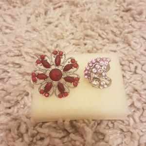 Två statementringar med stenar - Säljer två unika ringar: en stor blomformad ring med röda stenar och silverfärgad bas, samt en mindre ring dekorerad med rosa och klara stenar i en swirl-design. Perfekta för dig som gillar att sticka ut och addera färg till din stil. 5 kr/st.
