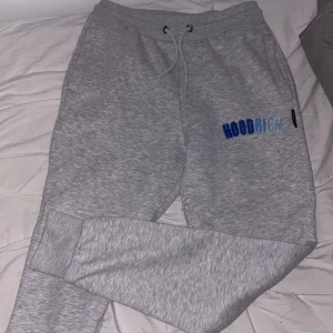 Grå mjukisbyxor från Hoodrich M - Snygga grå mjukisbyxor från Hoodrich i storlek medium. Byxorna har broderad blå logga på ena benet, dragsko i midjan och muddar vid bensluten. Perfekta för chill dagar eller när du vill ha en avslappnad stil.