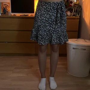 Svart blommig kjol från FB Sister - Supersöt svart kortkjol från FB Sister med vitt blommönster, volangkant och knyt i midjan. Kjolen har en flowy passform och är perfekt för vår och sommar. Tillverkad i lätt polyester som känns skön mot huden.