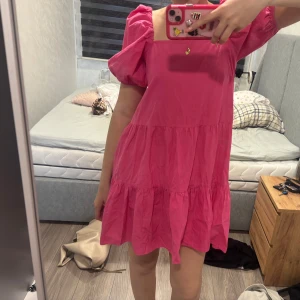 Rosa puffärmad kortklänning - Supersöt rosa klänning med puffärmar och volangdetaljer. Den har en fyrkantig urringning och är kort i modellen, perfekt för en trendig look. Lätt och luftig, passar perfekt till varma dagar.