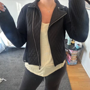 Svart quiltad jacka från Barbour - Säljer en svart jacka från Barbour med quiltat mönster framtill och ribbstickade ärmar. Jackan har sned dragkedja och en snygg, figurnära passform. 