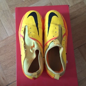 Nike Phantom GT fotbollsskor gul/orange - Nike Phantom GT fotbollsskor i en riktigt fet gul och orange färg med svarta detaljer och stora Swoosh-loggor. Skorna har låg profil, plös med hål för snörning och greppig sula med dobbar för gräsplan. Perfekta för dig som vill sticka ut på planen. Skorna är lite smutsiga men har ej testat att tvätta. Snöre ingår.