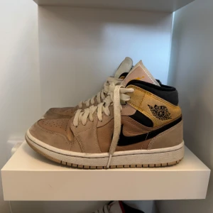 Nike Air Jordan 1 Mid beige/gul 37.5 - Säljer ett par Nike Air Jordan 1 Mid i beige mocka och läder med svarta detaljer och gul panel vid ankeln. Klassisk svart swoosh och vita snören. Perfekta för dig som gillar streetstyle och vill sticka ut med snygga färgkombos.