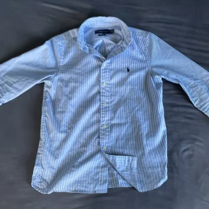 Blåvit randig skjorta Ralph Lauren - Snygg blå och vit randig skjorta från Ralph Lauren i slim fit-modell. Skjortan har klassisk krage, långa ärmar och knäppning framtill. Tillverkad i mjuk bomull och pryds av den ikoniska broderade loggan på bröstet. Perfekt för en clean och stilren look.