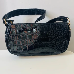 Svart axelväska i croco-look  - Snygg svart axelväska från SHEIN med glansig croco-imitation och guldfärgade detaljer. Väskan har en bred axelrem och dragkedja upptill. Perfekt accessoar för att lyfta din outfit med lite extra edge.