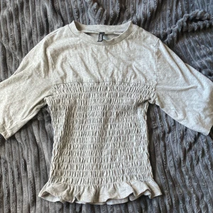 Grå topp med smock från H&M - Superfin ljusgrå topp från H&M med smockad midja och volangkant nertill. T-shirten har halvlånga ärmar och rund halsringning. Mjuk och skön i bomullsmix, perfekt för en chill och stilren look.