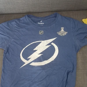 Blå Tampa Bay Lightning t-shirt Fanatics - Blå t-shirt från Fanatics med Tampa Bay Lightning-logga och Stanley Cup Champions-tryck fram. På ryggen står det Kucherov och nummer 86 i vitt. Skön bomull, perfekt för hockeyfans som vill visa sitt stöd.