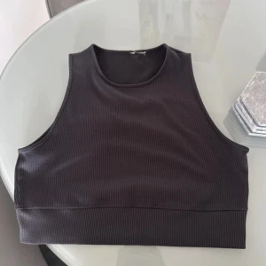 Svart ribbad linnetopp - En svart ribbad linnetopp med hög halsringning och ärmlös design. Toppen är croppad och har en tight passform som sitter snyggt mot kroppen. Perfekt att styla med jeans eller kjol för en clean look.