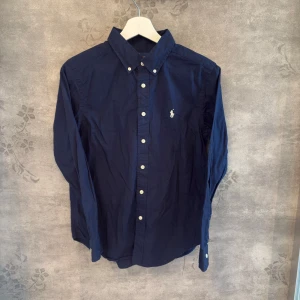 Polo Ralph Lauren, Skjorta Navy - 👔 Klassisk mörkblå skjorta från Polo Ralph Lauren 🔹 Storlek: L (14 - 16) 🔹 Färg: Djup navy blå, vit broderad logga  ✅ Äkta vara – köpt i butik 🧼 Nytvättad & fräsch – redo att användas direkt. Säljer den då den inte passade mig.