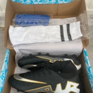 Svarta Nike fotbollsskor med guld - Snygga svarta Nike fotbollsskor med guld- och silverdetaljer samt tydlig AIR-logga på sidan. Skorna har snörning, platt sula med dobbar och en strumpliknande mudd runt vristen för extra support. Perfekta för dig som vill sticka ut på planen.