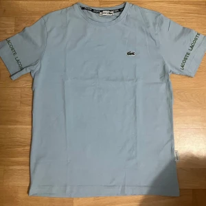 Ljusblå Lacoste t-shirt - Ljusblå t-shirt från Lacoste med klassisk krokodillogga på bröstet och broderad Lacoste-text på båda ärmarna. T-shirten har rund hals och är i mjuk bomull, perfekt för en clean och stilren look.