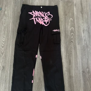 Minus Two svarta cargopants med tryck - Svarta cargopants från Minus Two med stora rosa graffiti-prints fram och bak. Byxorna har flera fickor på sidorna och bak, samt coola detaljer med rosa logga på benfickan. Perfekta för dig som gillar streetwear och vill sticka ut.