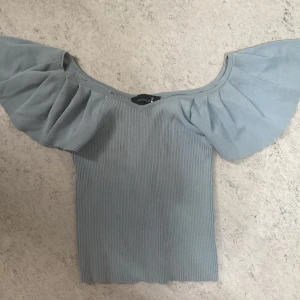 Ljusblå offshoulder topp med volangärm - Supersöt ljusblå topp från Jeanswest med ribbad kropp och stora volangärmar. Offshoulder-modell som ger en trendig vibe och passar perfekt till jeans eller kjol. Toppens material känns mjukt och stretchigt för en bekväm passform.