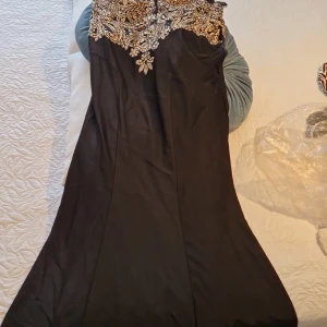 Svart långklänning med guldpaljetter - Elegant svart långklänning med fantastiska guldfärgade paljetter och broderier upptill. Klänningen har korta ärmar i transparent mesh och en figurnära passform som ger en lyxig känsla. Perfekt för bal eller fest där du vill sticka ut.