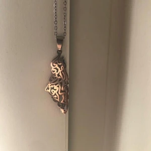 Unikt halsband i silver med mönster - Säljer ett halsband i silverfärg med ett coolt, abstrakt mönster i hänget. Kedjan är tunn och smidig, och hänget har en konstnärlig, nästan arabesk design som verkligen sticker ut. Perfekt för dig som gillar unika smycken med personlig touch.