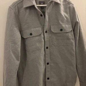 Grå overshirt från H&M - Stilren grå overshirt från H&M med klassisk krage och svarta knappar framtill. Två stora bröstfickor med lock och knapp ger en cool utility-vibe. Perfekt lager-på-lager-plagg med lång ärm och rak passform.