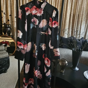 Zara - Långklänning med blommor - Supersnygg svart långklänning med stora blommor i rosa, rött och beige. Klänningen har hög krage, långa ärmar och dekorativa knappar framtill. Materialet känns lätt och glansigt, vilket ger en lyxig känsla. Perfekt för dig som vill sticka ut med en elegant look.