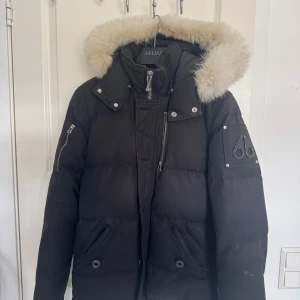 Svart dunjacka Moose Knuckles med päls - Säljer en svart dunjacka från Moose Knuckles med fluffig ljus päls runt huvan. Jackan har flera dragkedjor, tryckknappar och ikonisk logga på ärmen. Perfekt för kalla vinterdagar och har en riktigt snygg streetstil. Det är storlek S men passar M och den har sett bättre dagar därav priset 