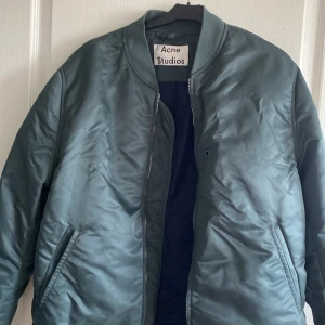 Grön bomberjacka från Acne Studios - Snygg grön bomberjacka från Acne Studios med klassisk ribbad krage och ärmslut. Jackan har dragkedja framtill och två snedställda fickor. Materialet är glansigt och känns riktigt modernt. Perfekt för dig som vill ha en stilren och trendig look.