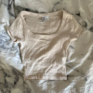 Beige ribbad topp från H&M Divided  - Säljer en beige ribbad topp från H&M Divided i storlek XS. Toppen har korta ärmar, rundad djup urringning och croppad passform. Perfekt att matcha med jeans eller kjol för en enkel och snygg look.