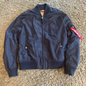 Mörkblå bomberjacka Alpha Industries - Snygg mörkblå bomberjacka från Alpha Industries med klassisk passform. Jackan har dragkedja framtill, ribbade muddar och fickor med tryckknappar. Detalj med röd tag på ärmen ger en cool touch. Säljer den då den är för liten.