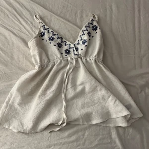 Vit broderad linnetopp från Shein - Supersöt vit linnetopp från Shein med blå broderade blommor och v-ringning. Toppen har smala axelband, knyt i midjan och en flowy passform. Perfekt för varma dagar och ger en fräsch och somrig vibe.
