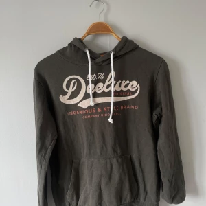 Mörkgrön hoodie från Deeluxe74 - Snygg mörkgrön hoodie från Deeluxe74 med vit tryckt logga och text på bröstet! Bra skick och använd fåtal gånger, storlek S men passar även XS!