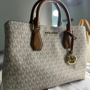 Beige handväska från Michael Kors - Snygg beige handväska från Michael Kors med MK-logga över hela väskan. Väskan har bruna handtag i skinn och ett guldigt MK-hänge. Perfekt storlek för allt du behöver och riktigt stilren design med diskret mönster. Använd endast en gång.