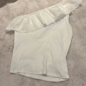 Vit offshoulder topp med volang Kappahl - Supersöt vit offshoulder topp från Kappahl med bred volangkant i broderad spets. Toppen är ärmlös och har en snygg asymmetrisk axel. Perfekt för dig som gillar romantisk och trendig stil.