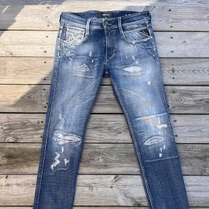 Replay Anbass blå slitna jeans - Säljer ett par Replay Anbass jeans i blå tvätt med slitningar och distressed detaljer. Jeansen har klassisk femficksdesign, smal passform och snygga kontrastsömmar. Perfekta för dig som gillar en trendig och avslappnad look.