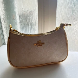 Beige axelväska från Coach - Snygg beige axelväska från Coach med guldiga detaljer och dragkedja. Väskan har ett subtilt mönster i materialet och kommer med en avtagbar axelrem. Perfekt storlek för det viktigaste och har en klassisk, stilren look. AA copy