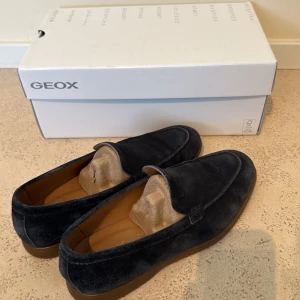 Marinblå mockaloafers från Geox - Snygga marinblå loafers från Geox i mjuk mocka med klassisk design och platt sula. Skorna har en stilren look och passar perfekt till både jeans och chinos. Bekväm innersula och diskret branding på sulan. Perfekta för dig som gillar en clean och tidlös stil. Köpa från zalando för något år sedan och lite använda. Nypris runt 1500kr. Hör av er vid funderingar! 