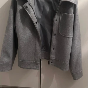 Grå overshirt med stor ficka - Snygg grå overshirt med klassisk krage och stor bröstficka. Jackan har raka linjer, knappar framtill och är tillverkad i ett mjukt ullmaterial. Perfekt för lager-på-lager och ger en clean look till din outfit.