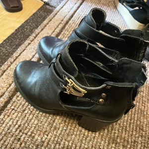 Svarta boots med spänne och klack - Snygga svarta boots med chunky klack, dekorativa guldfärgade spännen och coola cut-outs vid ankeln. Dragkedja baktill för enkel på- och avtagning. Gjorda i skinnimitation och passar perfekt till både jeans och kjol.