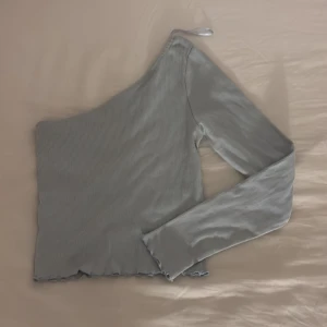 Ljusgrå ribbad oneshoulder topp - Snygg blå ribbad topp med oneshoulder-design och lång ärm. Toppen har en asymmetrisk form.