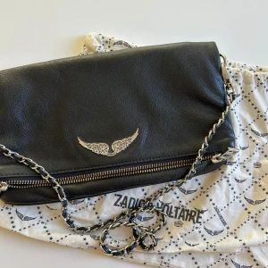 Zadig & Voltaire väska - Zadig & Voltaire Rock Clutch väska. Svart kuvertväska i läder med dubbla läder- och metallkedjor samt strassprydda vingar. Kan bäras på flera sätt med ett kortare och ett längre band.   Väskan har stängning med dragkedja, magnetisk stängning, liten bakficka med dragkedja och innerficka.  Mått: 27 x 13 x 2 cm  Säljes pga kommer inte till användning.