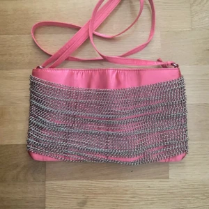 Rosa axelväska med kedjedetaljer Gina Tricot - Snygg rosa axelväska från Gina Tricot i glansigt satinliknande material. Väskan har coola silverfärgade kedjor på framsidan och en lång axelrem. Perfekt för dig som vill sticka ut med en unik accessoar.