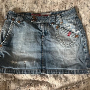 Ljusblå jeanskjol från Anna Koola - Snygg ljusblå jeanskjol från Anna Koola med slitna detaljer och klassiska fickor fram och bak. Kjolen har bälteshällor, knappar i metall och en cool tvättad look. Perfekt för dig som gillar en avslappnad och trendig stil.