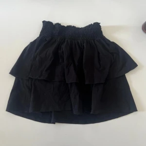 Svart volangkjol från Gina Tricot - Supersöt svart kortkjol från Gina Tricot med volanger och smockad midja. Kjolen är gjord i mjuk bomull och har en flowy känsla som ger en chill och trendig vibe. Perfekt att styla med en crop top eller hoodie.