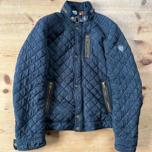 Mörkblå jacka från Saki - Quiltad mörkblå jacka från Saki med detaljer som dragkedjor, knappar och spänne vid kragen. Jackan har lång ärm, ståkrage och flera fickor med dragkedja. Ett litet håll finns på framsidan. Går givetvis att diskutera priset.