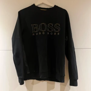 Svart sweatshirt från Hugo Boss - Svart sweatshirt från Hugo Boss med stor BOSS-logga framtill i kontrasterande färg. Klassisk rund halsringning och långa ärmar. Perfekt för en clean och stilren look.