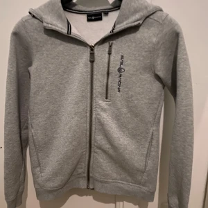 Grå Sail Racing hoodie med dragkedja - Grå hoodie från Sail Racing med broderad logga på bröstet och huva. Tröjan har hel dragkedja framtill, en extra bröstficka med dragkedja och långa ärmar. Köpt för 1199 kr på kidsbrandstore och säljs för 499 kr. Endast hämtning som gäller. Hör av er vid frågor gällande tröjan.