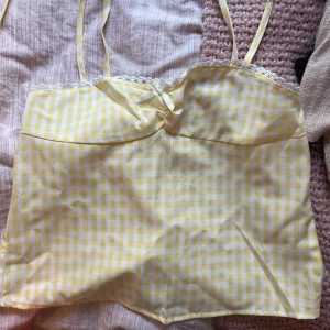 Gul och vit rutig linnetopp med rosett - Supersöt linnetopp i gulvitt rutigt mönster med tunna axelband, liten rosett framtill och spetskant upptill ifrån shein. 