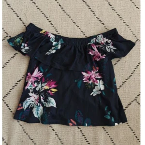 Blommig offshoulder topp - Snygg offshoulder topp med stora blommiga mönster i rosa, vitt och grönt. Toppen har volang upptill och är ärmlös, perfekt för varma dagar. Lätt och luftig känsla, passar till jeans eller kjol.