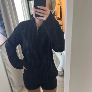 Svart ribbad stickad tröja half zip - Svart stickad tröja med ribbad struktur och half zip-dragkedja i halsen. Tröjan har en avslappnad passform och långa ärmar, perfekt för lager på lager under kyliga dagar. Enkel och stilren design som funkar till det mesta.