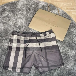 Burberry shorts - Snygga badshorts från Burberry i klassiskt rutigt mönster i grått, svart och beige. Shortsen har elastisk midja med svart snörning och innerbyxa i mesh. Perfekta för stranden eller poolen, riktigt stilrena och ikoniska.