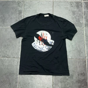 Svart Moncler t-shirt med stor logga - Svart t-shirt från Moncler med stor, grå och vit logga framtill. Loggan har broderade detaljer i rött och blått som ger en cool kontrast. Klassisk passform och rund halsringning. Perfekt för dig som gillar streetwear och exklusiva märken. Storleken är S men passar även M! Hör av dig vid frågor eller funderingar!🤝🏻
